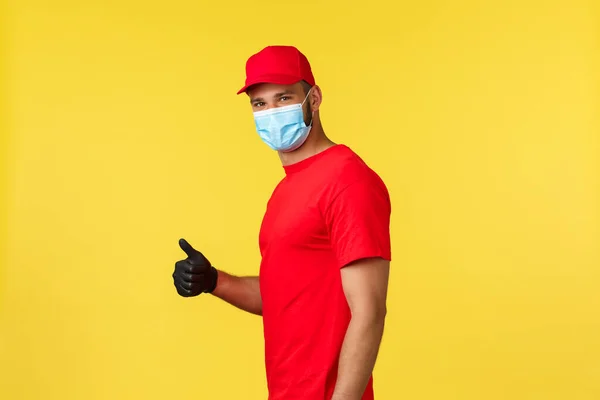 Express delivery during pandemic, covid-19, safe shipping, online shopping concept. Smiling handsome courier in red uniform and medical mask, show thumb-up, recommend package deliver service — Stock Photo, Image