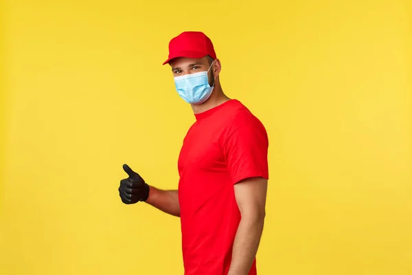 Consegna espressa durante pandemia, covid-19, spedizione sicura, concetto di shopping online. Sorridente corriere bello in uniforme rossa e maschera medica, mostrare pollice in su, raccomandare pacchetto consegnare servizio — Foto Stock