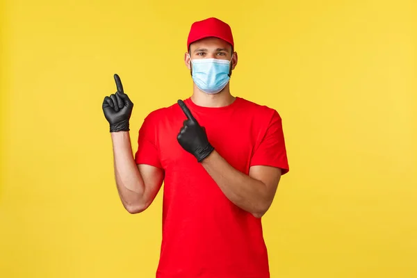 Entrega expressa durante pandemia, covid-19, transporte seguro, conceito de compras on-line. Correio sorridente em máscara médica e tampa uniforme vermelho, t-shirt, apontando os dedos canto superior esquerdo, mostrar banner — Fotografia de Stock