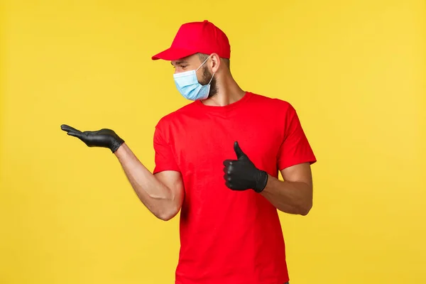 Express delivery during pandemic, covid-19, safe shipping, online shopping concept. Pleased courier in red uniform cap, t-shirt and medical mask show thumb-up and looking at palm satisfied — Stock Photo, Image