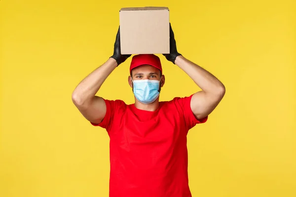 Express delivery during pandemic, covid-19, safe shipping, online shopping concept. Smiling enthusiastic courier in red uniform, medical mask and gloves, holding box package on head — Stock Photo, Image