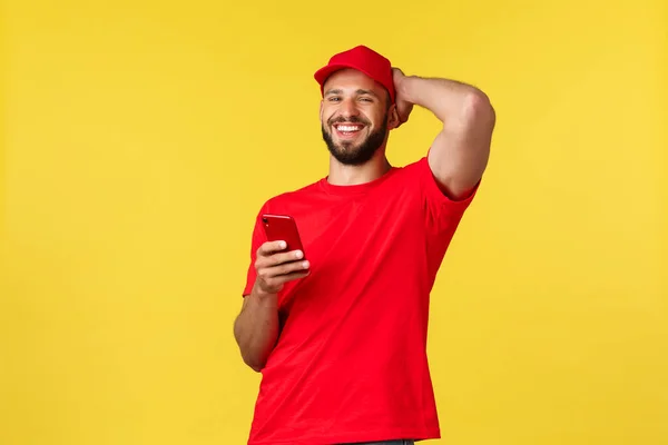 无接触支付，网上交货，网上购物和包裹跟踪的概念。无忧无虑的小胡子小伙子，戴着红帽子和T恤的信使，笑得像在用手机，黄色背景 — 图库照片
