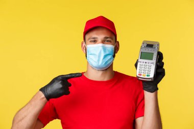 Covid-19, teslimat emirleri, alışveriş, temassız ödeme ve sosyal uzaklık konsepti. Yakından gülümseyen, kırmızı üniformalı, tıbbi maskeli, eldivenli, ödeme terminalini gösteren yakışıklı bir kurye.