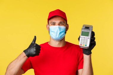 Covid-19, teslimat emirleri, alışveriş, temassız ödeme ve sosyal uzaklık konsepti. Kırmızı üniformalı tatmin olmuş yakışıklı kurye, koronavirüs salgını sırasında güvenlik için POS terminalini kullan