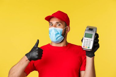 Covid-19, teslimat emirleri, alışveriş, temassız ödeme ve sosyal uzaklık konsepti. Kırmızı üniformalı, tıbbi maske ve eldivenli etkileyici ve heyecanlı kurye, baş parmağını kaldır ve POS terminalini göster.