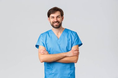 Sağlık çalışanları, koronavirüs karantina kampanyası ve salgın konsepti. Gülümseyen sakallı beyaz erkek doktor ya da mavi önlüklü hemşire, çapraz göğüslü ve gülüyor.