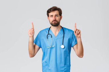 Sağlık çalışanları, salgın hastalık ve koronavirüs salgını konsepti. Üzgün, tedirgin, sakallı doktor önlüklü, tıbbi giysili ve steteskoplu, parmaklarını havaya kaldırmış, hayal kırıklığına uğramış ve yorgun.