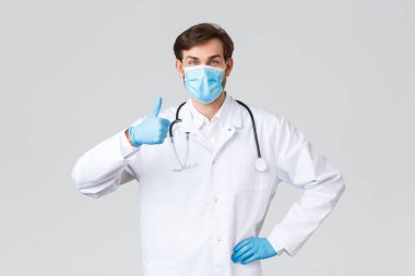 Hastane, sağlık çalışanları, covid-19 tedavi konsepti. Tıbbi maskeli, önlüklü ve eldivenli iyimser destekçi doktor, fikir gibi onaylanmış, hasta neşelendirme, gri arka plan