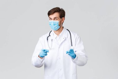 Hastane, sağlık çalışanları, covid-19 tedavi konsepti. İyi gidiyorsun. Tıbbi maskeli neşeli doktor ve eldivenli önlüklü, kamerayı işaret eden, hastayı selamlayan, gri arka planlı