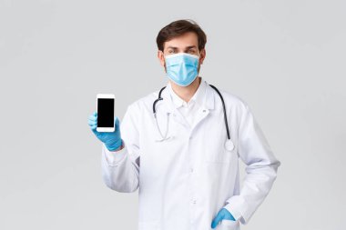Hastane, sağlık çalışanları, covid-19 tedavi konsepti. Tıbbi maske takan gülümseyen yakışıklı doktor, ameliyat önlüğü ve eldivenleri hastaların kendi kendilerini karantinaya alırken iletişim kliniğine akıllı telefon başvurularını gösteriyorlar.