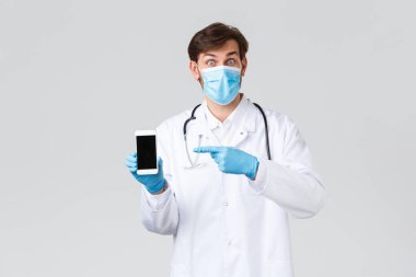 Hastane, sağlık çalışanları, covid-19 tedavi konsepti. Tıbbi maskeli, eldivenli, önlüklü, akıllı telefonu işaret eden şaşkın doktor internette harika bir tanıtım ve iyi bir uygulama buldu.