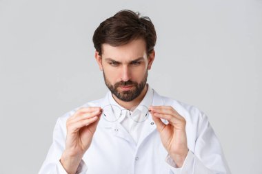 Hastane, sağlık çalışanları, covid-19 tedavi konsepti. Gözlüklü ciddi görünümlü bir doktor, sağ optik lensli, ayakta duran gri arka planı olan hastaya gözlük reçetesi yazıyor.