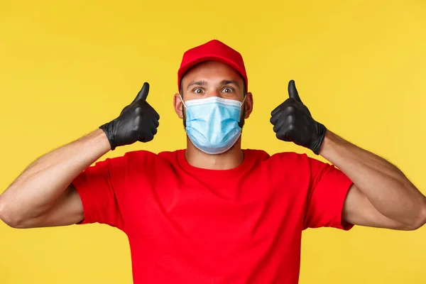 Covid-19, delivery orders, shopping, contactless payment and social distancing concept. Upbeat and excited male courier in red uniform, medical mask, show thumbs-up in approval — Stock Photo, Image