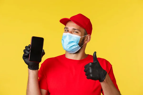 Covid-19, delivery orders, shopping, contactless payment and social distancing concept. Handsome male courier in red uniform, medical mask and gloves, advice download application for order tracking