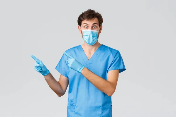 Covid-19, vårdpersonal och sjukhuskoncept. Förvånad och upphetsad läkare i medicinsk mask och sjuksköterska kostym, pekande fingrar vänster, stirrande förvånad. Praktikant ställa frågan visar vägen — Stockfoto