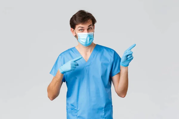 Covid-19, trabajadores sanitarios y concepto de hospital. Doctor invitando a mirar la publicidad, mira. Enfermera en máscara médica, guantes y exfoliantes apuntando con los dedos a la derecha, mostrando promo, fondo gris —  Fotos de Stock