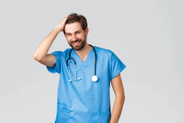 Covid-19, kwarantanna, szpitale i opieka zdrowotna. Przystojny brodaty lekarz lub pielęgniarka w niebieskim fartuchu ze stetoskopem, biegająca ręka przez włosy i uśmiechnięta kamera — Zdjęcie stockowe