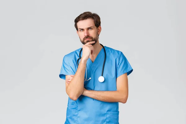 Covid-19, quarantine, hospitals and healthcare workers concept. Thoughtful doctor in scrubs, stethoscope, rubbing chin and frowning, thinking serious, solving problem, look at patient — Stock Photo, Image