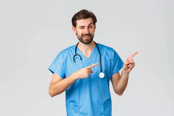 Healthcare workers, pandemic and coronavirus outbreak concept. Handsome cheeky doctor in medical scrubs, stethoscope, pointing fingers right and wink smile, showing way or promo — Stock Photo, Image