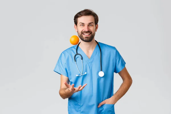 Gezondheidswerkers, pandemie en coronavirus uitbraak concept. Knappe chirurg in scrubs en stethoscoop, dokter flipping oranje en glimlachen, vertellen patiënten eten vitaminen en fruit — Stockfoto