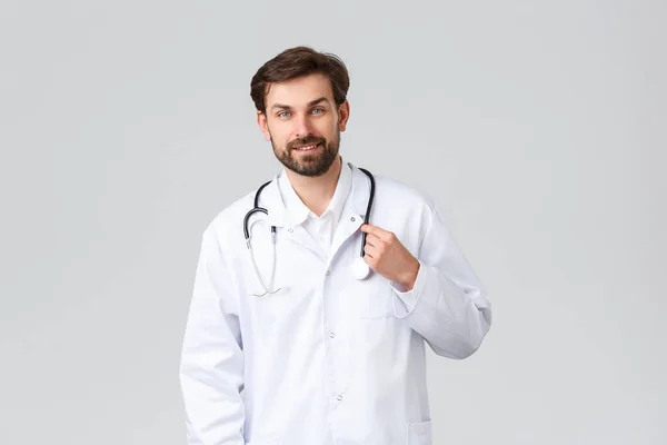 Hôpital, travailleurs de la santé, concept de traitement covid-19. Beau docteur barbu en blouse blanche avec stéthoscope, souriant au patient, écoutant les problèmes, traitant les gens à la clinique — Photo