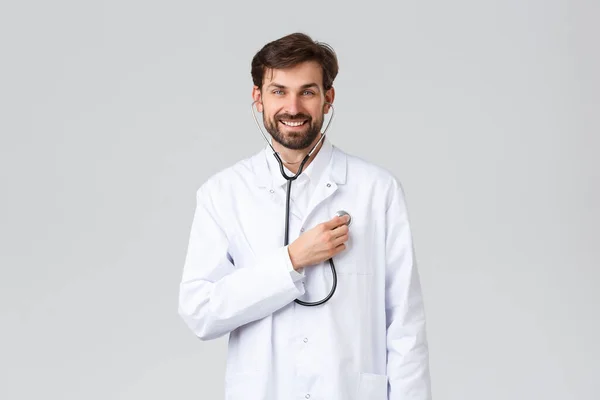 Hospital, healthcare workers, covid-19 treatment concept. Handsome smiling bearded doctor in white scrubs, wearing stethoscope and listen to his heartbeat, physician working in clinic — Stock Photo, Image