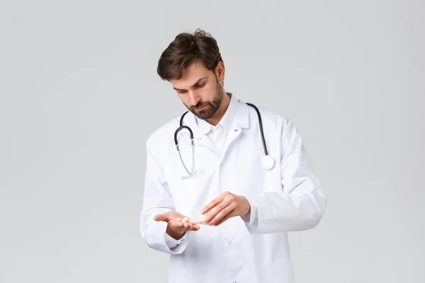 Больница, медицинские работники, концепция лечения ковид-19. Серьёзно выглядящий доктор в белом халате со стетоскопом, подсчитывающий таблетки, лекарства в руке, выглядящий обеспокоенным или решительным — стоковое фото