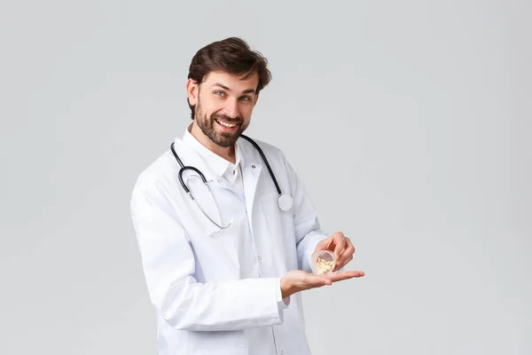 Hospital, healthcare workers, covid-19 treatment concept. Handsome bearded physician or doctor in white scrubs with stethoscope, showing pills, promo of medication, prescribed drugs — Stock Photo, Image