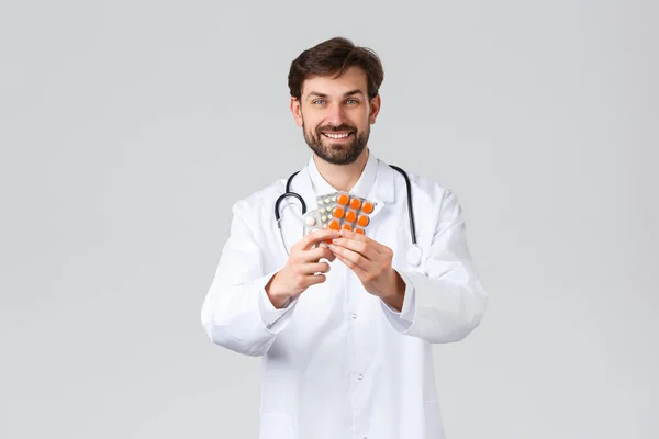 Szpital, pracownicy służby zdrowia, koncepcja leczenia. Uśmiechnięty, przystojny lekarz w białym fartuchu ze stetoskopem, pokazujący leki, rekomendujący tabletki lub witaminy, szare tło — Zdjęcie stockowe