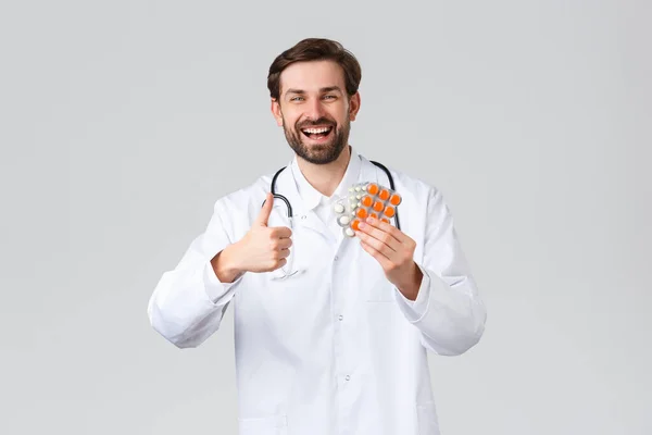 Больница, медицинские работники, концепция лечения ковид-19. Счастливый веселый белый врач улыбается, показать большой палец вверх в утверждении и советы лекарства, витамины или таблетки, способствовать здравоохранению — стоковое фото