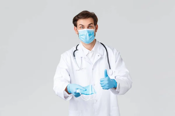 Hastane, sağlık çalışanları, covid-19 tedavi konsepti. Önlüklü neşeli bir doktor, koronavirüs sırasında koruyucu ekipman, tıbbi maske takmanın gerekliliğini açıklıyor, baş parmağını kaldırıyor. — Stok fotoğraf