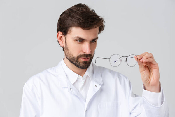 Hospital, healthcare workers, covid-19 treatment concept. Close-up handsome bearded doctor in scrubs, looking closely to glasses lense, picking appropriate lense for eyewear, grey background
