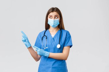 Covid-19, virüsün önlenmesi, sağlık, sağlık çalışanları ve karantina konsepti. Şaşırmış kadın hemşire ya da doktor mavi önlüklü, tıbbi maske ve eldivenli, sola bakıyor ve onu işaret ediyor.
