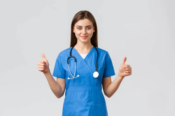 Healthcare workers, prevent virus, insurance and medicine concept. Supportive professional female nurse or doctor in blue scrubs, stethoscope, show thumbs-up in approval, smiling — Stock Photo, Image