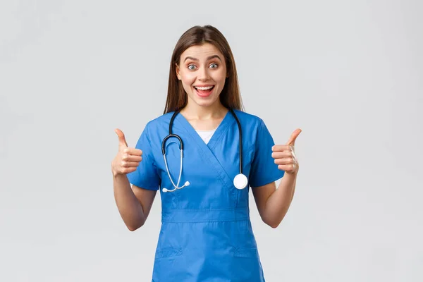 Sjukvårdspersonal, sjukvård, covid-19 och vaccinationskoncept. Entusiastiska och optimistiska kvinnliga sjuksköterska, läkare i blå skurar, tycker underbar idé, visa tummen upp i godkännande — Stockfoto