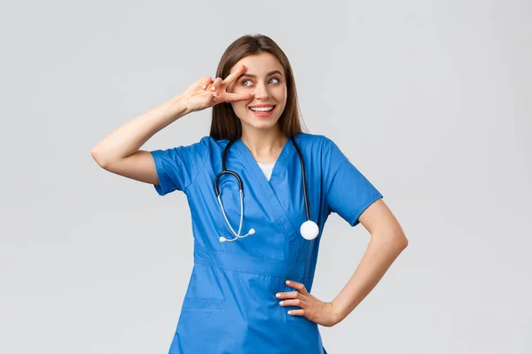 Pracownicy medyczni, opieka zdrowotna, koncepcja covid-19 i szczepień. Wesoły i energiczny młody lekarz, ładna pielęgniarka w niebieskim fartuchu, stetoskop, pokazać spokój w pobliżu oka i uśmiechnięty optymizm — Zdjęcie stockowe