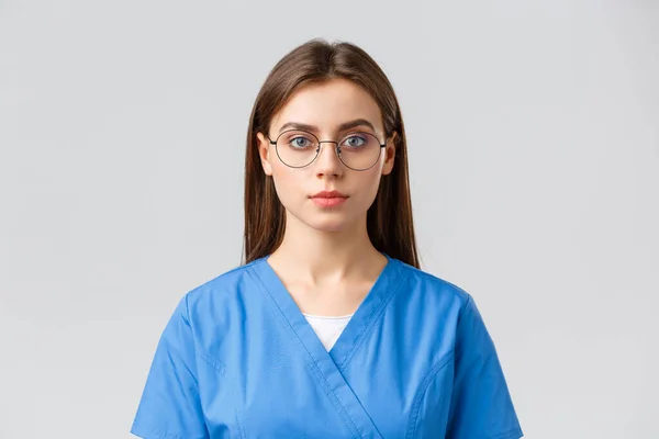 Egészségügyi dolgozók, orvostudomány, biztosítás és a kovid-19 világjárvány koncepciója. Fiatal női gyakornok, ápolónő vagy orvos műtősruhában és szemüvegben, komoly arckifejezéssel, szürke háttérrel nézi a kamerát — Stock Fotó
