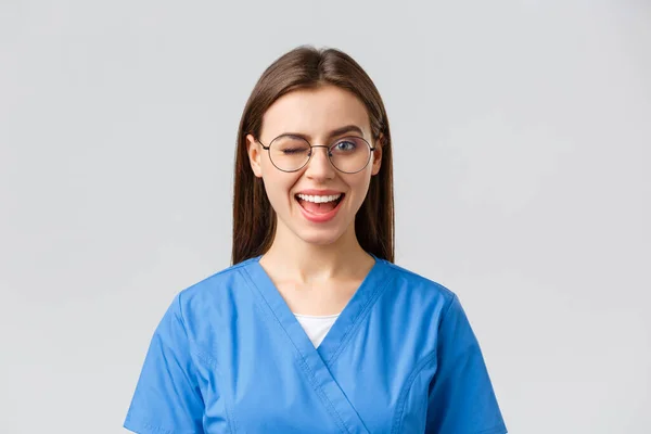 Sjukvårdspersonal, medicin, försäkring och covid-19 pandemi koncept. Fräcka söta kvinnliga sjuksköterska, praktikant eller läkare på kliniken, bära skurar och glasögon, blinka vid kameran med entusiastiskt leende — Stockfoto