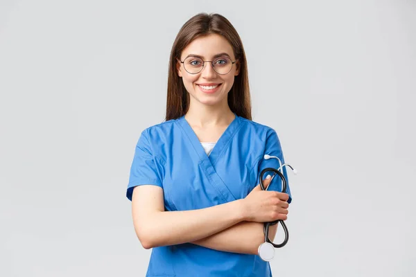 Gesundheitspersonal, Medizin, Versicherung und Covid-19-Pandemiekonzept. Selbstbewusste und begeisterte junge Krankenschwester, Ärztin in blauem Peeling und Brille, Stethoskop in der Hand, lächelnde Kamera erfreut — Stockfoto