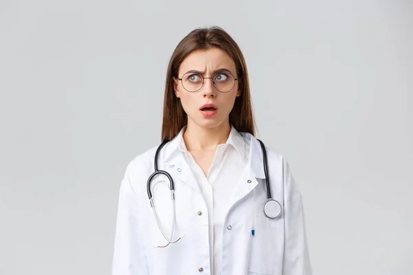 Trabajadores sanitarios, medicina, seguros y concepto de pandemia covid-19. Doctora joven conmocionada y confundida en uniformes blancos y anteojos, estetoscopio, mirada fija en la cara nerviosa preocupada — Foto de Stock