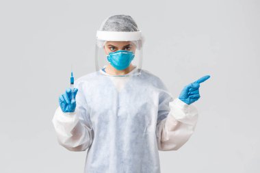 Covid-19, tıbbi araştırma, teşhis, sağlık çalışanları ve karantina konsepti. Kişisel koruyucu kostümlü ciddi bir doktor, tıbbi solunum cihazı, solu işaret ediyor, şırıngayı aşıyla tutun.