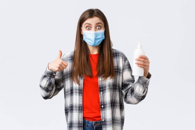 Coronavirus salgını, karantina, sosyal mesafeler ve duygu kavramı. Tıbbi maskeli heyecanlı ve tatmin olmuş bir kız el dezenfektanı, virüs sabununu ve parmak uçlarını önlüyor.
