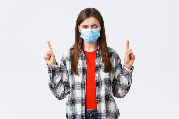 Coronavirus uitbraak, vrije tijd op quarantaine, sociale afstand en emoties concept. Boze, verontwaardigde jonge vrouw met medisch masker grimmig, die er gek uitziet alsof ze naar boven wijst. — Stockfoto