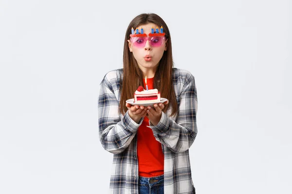 People lifestyle, holidays and celebration, emotions concept. Cute tender young b-day girl in glasses, blowing lit candle on birthday cake, making wish and celebrating at party, white background