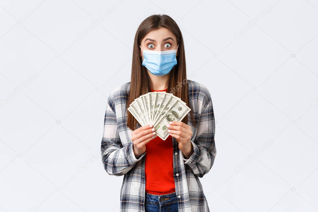 Money transfer, investment, covid-19 pandemic and working from home concept. Excited happy woman in medical mask received first paycheck, showing cash and look astonished