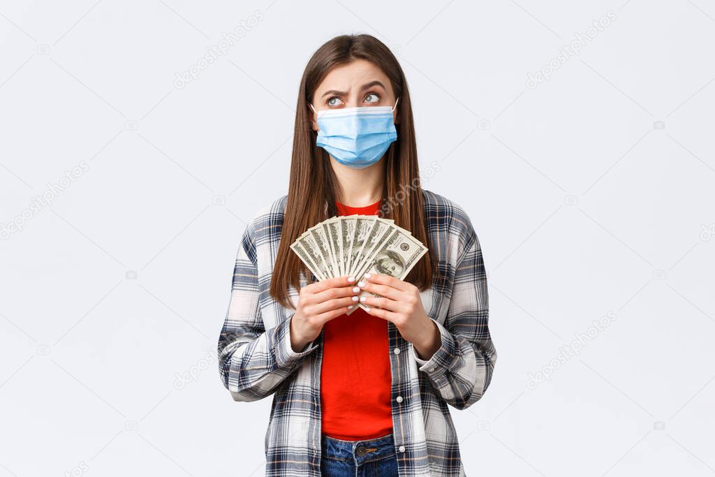 Money transfer, investment, covid-19 pandemic and working from home concept. Thoughtful and concerned woman in medical mask, look away and hold cash, thinking how save