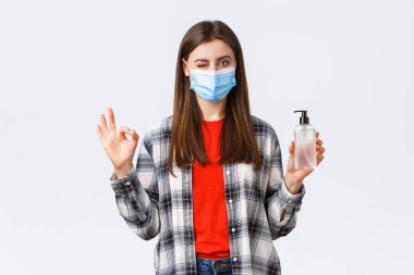 Coronavirus salgını, karantina, sosyal mesafeler ve duygu kavramı. Hiç sorun değil. Tıbbi maskeli hoş kız el dezenfektanı gibi işaret ve göz kırp.