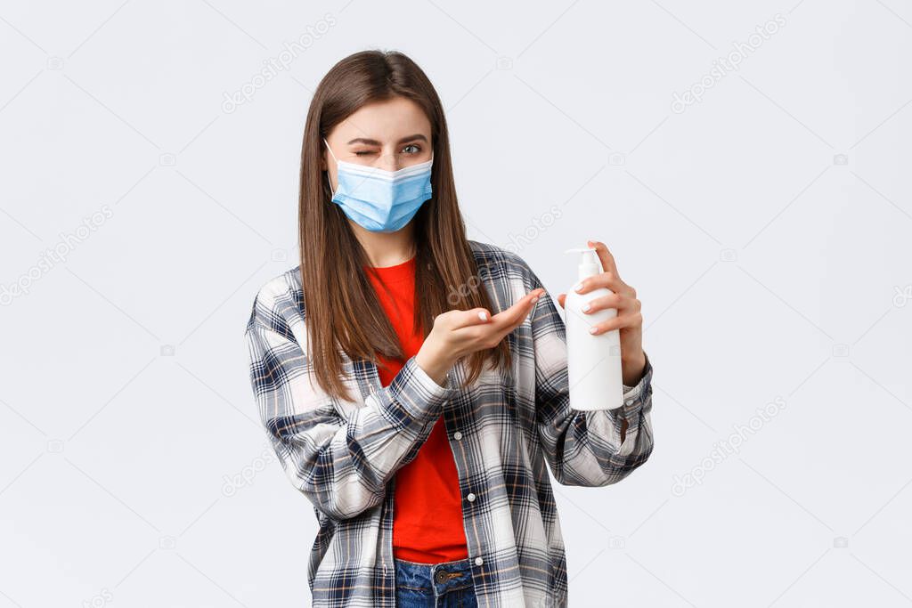 Coronavirus outbreak, leisure on quarantine, social distancing and emotions concept. Cheeky cute woman in medical mask, wink at you as apply hand sanitizer or soap, preventing virus