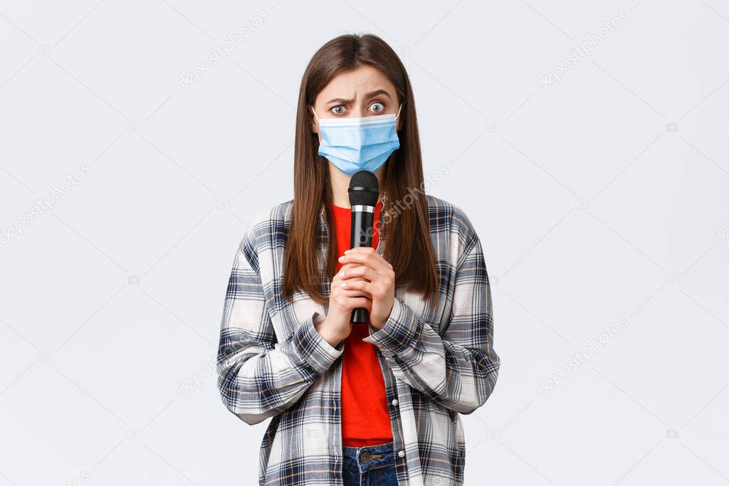 Coronavirus outbreak, leisure on quarantine, social distancing and emotions concept. Confused woman in medical mask look smth strange, holding microphone, perform, white background