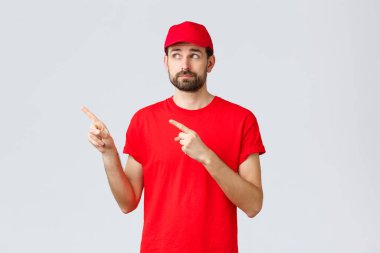 İnternetten alışveriş, karantina sırasında teslimat ve paket servis konsepti. Belirsiz ve hoşnutsuz, isteksiz kurye, kırmızı tişörtlü ve şapkalı çalışan, sol üst köşedeki parmaklarını göstererek hayal kırıklığına uğramış sırıtış.